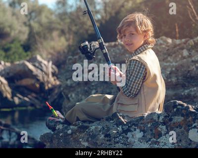 Side view of boy looking at camera delightfully and holding fishing rod  while sitting on rocky coast in selective focus Stock Photo - Alamy