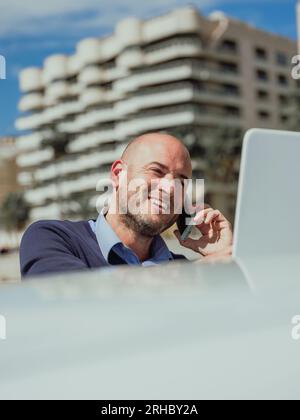 Stock photo of a businessman talking on the phone while using the laptop supported by a car with the buildings of the city behind. Lifestyle Stock Photo