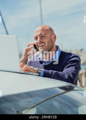 Stock vertical photo of a businessman talking on the phone while using a laptop supported by a car Stock Photo
