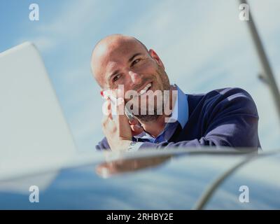 Stock photo of businessman talking on the phone while using the laptop supported by a car and smiling expression. Lifestyle Stock Photo