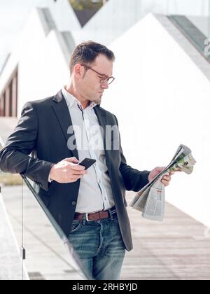 Serious male entrepreneur in eyeglasses browsing cellphone while reading newspaper near glass fence on sunny street with building in city Stock Photo