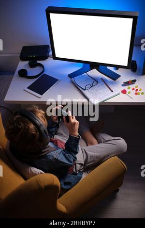 High angle of anonymous child in headset sitting with joystick in armchair while playing video game in dark room Stock Photo