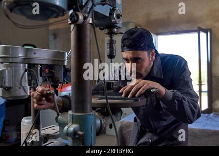 Concentrated adult bearded Hispanic male technician carefully using vertical drilling machine in workshop with opened door at daytime Stock Photo