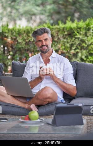 Bearded male freelancer with gray hair smiling and looking away while drinking coffee and working remotely on laptop in courtyard Stock Photo