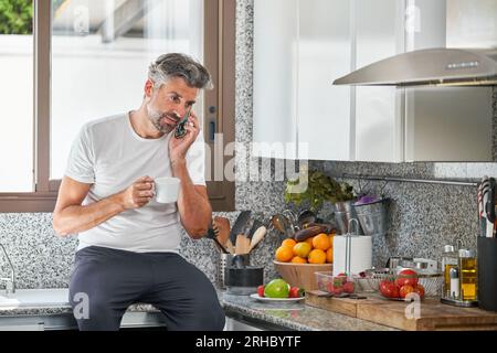 Mature bearded man in white t shirt sitting on counter near window and making phone call while drinking aromatic coffee in morning in kitchen Stock Photo