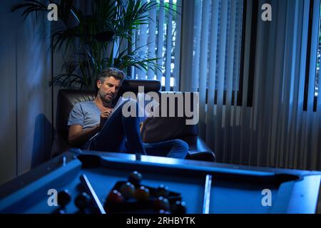 Mature male employee in casuals reading documents on chair working late in recreation room at office Stock Photo