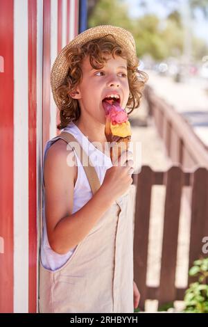 Side view of happy kid in overall and straw hat licking refreshing ice cream and looking away while standing near striped wall on city street on summe Stock Photo