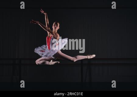 Full body of graceful female ballerina in costume and tutu and pointe shoes dancing with raised hands while dancing ballet role in rehearsal in dark s Stock Photo