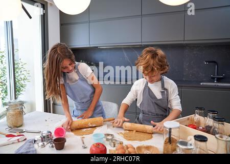Happy cute little siblings with blond hair in casual clothes and aprons smiling and kneading dough with wooden rolling pin while preparing cookies in Stock Photo