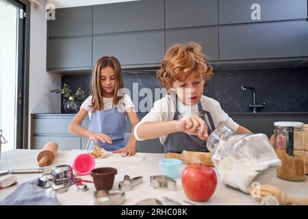 Adorable children in aprons making sweet dough for cookies with flour at table with various utensils in light modern kitchen Stock Photo