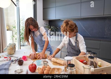 Smiling little girl and boy with blond hair in similar aprons kneading soft dough while preparing delicious homemade cookies in kitchen Stock Photo