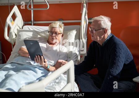 Side view of focused elderly man sitting near sick wife lying on medical bed and watching video on tablet during recovery process in modern hospital i Stock Photo