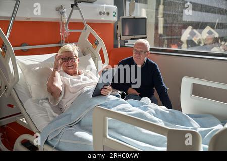 Happy senior female patient smiling and looking away while lying on medical couch with tablet and adjusting eyeglasses near husband during rehabilitat Stock Photo