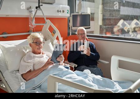 Elderly male browsing mobile phone while sitting against senior unwell wife lying on bed with tablet in modern hospital Stock Photo