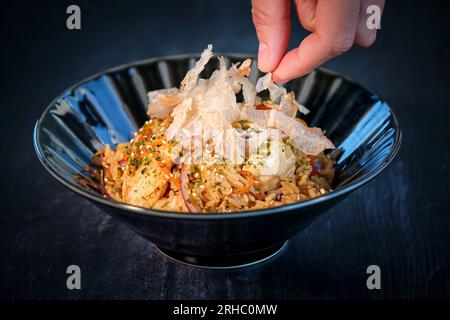 From above of anonymous crop person putting crisps on spicy fried rice with ingredients of pepper onions and tomatoes in bowl placed on black surface Stock Photo