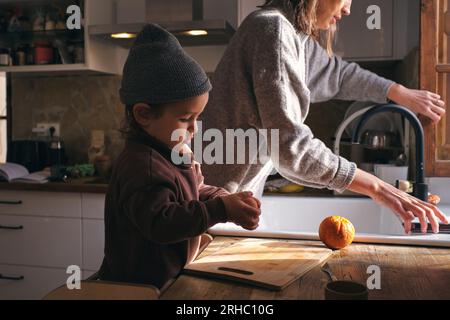 Side view of cute little child peeling oranges on cutting board standing at table near crop mother in kitchen Stock Photo