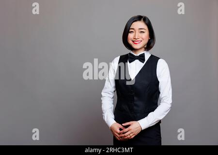 Smiling asian restaurant waitress wearing black and white uniform standing and looking at camera. Cheerful young woman professional receptionist posing for studio portrait Stock Photo