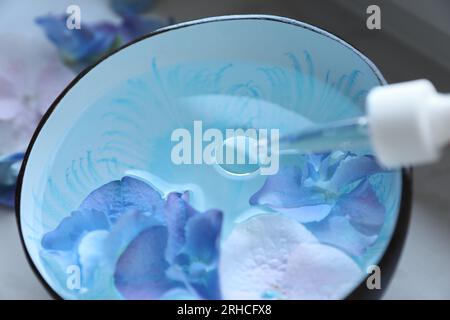 Dripping essential oil from pipette into bowl with water and flowers, closeup Stock Photo