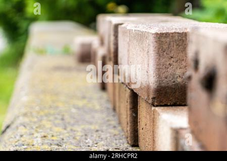 Close up of reddish-brown bricks on light gray concrete wall with moss - rough texture and staggered pattern - blurred garden background Stock Photo