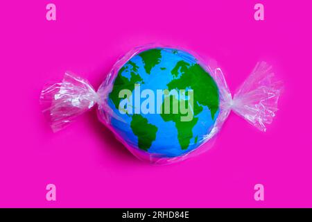Miniature globe wrapped in plastic packaging, resembling a wrapped candy, isolated on pink background. Plastic pollution related concept. Stock Photo