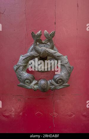 Closeup view of traditional vintage brass door knocker with stylized fish isolated on ancient dark red wooden door, Montpellier, France Stock Photo