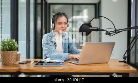 A beautiful Asian female podcaster or radio host working in her studio, speaking into a microphone, recording her podcast. Stock Photo