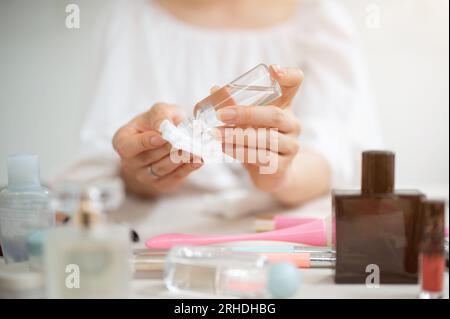 Close-up image of a woman using facial makeup remover, dropping makeup remover on a cotton pad to clean her face while sitting at her vanity desk. Stock Photo