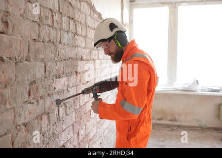Construction worker in orange coverall removing old plaster from red brick wall by hammer drill. Stock Photo