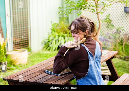 Happy young woman sitting at picnic bench in garden with pet lizards working on tablet laptop Stock Photo