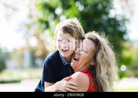 Grandmother carrying her grandson, laughing together outside at a park. she is his caregiver, he has autism Stock Photo