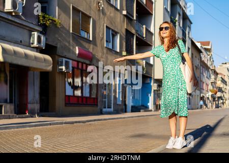 Young urban woman wearing summer dress and sunglasses hailing a taxi on the street. Stock Photo