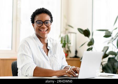 Nice and friendly female colleague looking at te camera with wide charming smile with gap teeth, african female entrepreneur, ceo, manager laughing and using laptop, excited happy business woman Stock Photo