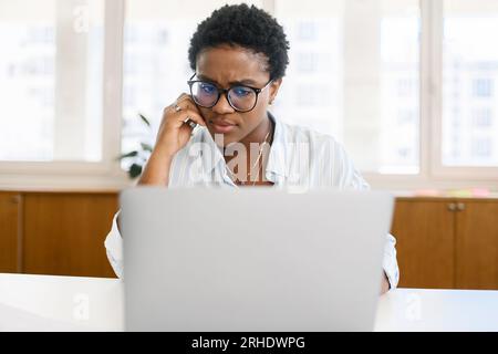 Puzzled pensive african-american female employee sitting at desk in front of laptop, thinking and feeling doubt. Serious freelancer woman trying to solve business problem, looking undistracted Stock Photo