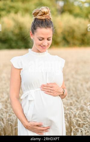 Smiling young prenatal female in white dress looking down while touching baby bump with hands and standing in grassy field on sunny day in countryside Stock Photo