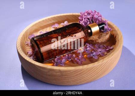 Bottle with essential oil, lilac flowers and water in bowl on violet background Stock Photo