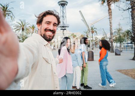 Handsome young adult man taking a selfie portrait holding his girlfriend's hand while they are walking in the city with their joyful friends. International people having fun together on weekend. High quality photo Stock Photo