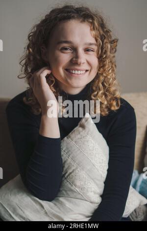 Morning portrait of smiling pretty young curly-haired woman smiling and looking at camera holding a pillow in her arms. Happiness and start of new day Stock Photo