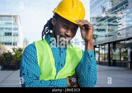 black man engineer builder, standing outdoors waving and smiling holding his hardhat with one hand, people concept, copy space. Stock Photo