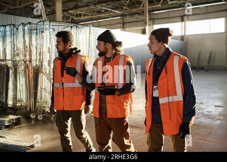 Three young intercultural builders in safety jackets walking along spacious workshop and discussing which new equipment should be acquired Stock Photo