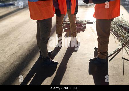 Cropped shot of three young male builders in flourescent jackets standing in front of camera in warehouse and communicating at meeting Stock Photo