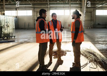 Group of three young intercultural builders in workwear discussing working points at meeting while standing in warehouse or workshop Stock Photo