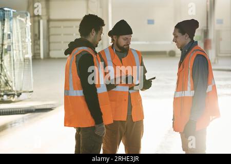 Two young multiethnic male engineers in protective flourescent vests listening to their foreman with tablet at meeting in factory warehouse Stock Photo