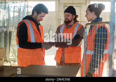 Three young male workers of warehouse in reflective jackets discussing list of delivered goods while standing by cart with stack of boxes Stock Photo