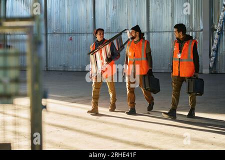 Three happy engineers of warehouse with stepladder and toolboxes looking at one another and having chat while walking forwards Stock Photo