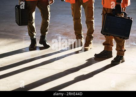Legs of three male engineers with toolboxes standing on sunlit floor of modern warehouse or industrial plant and having working meeting Stock Photo