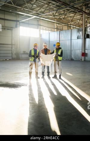 Long shot of three young builders or engineers in hardhats discussing blueprint with building plan while standing in sunlit warehouse Stock Photo