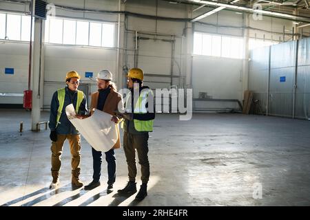 Long shot of two young multiethnic engineers in protective workwear and contractor with blueprint looking at building plan and consulting Stock Photo