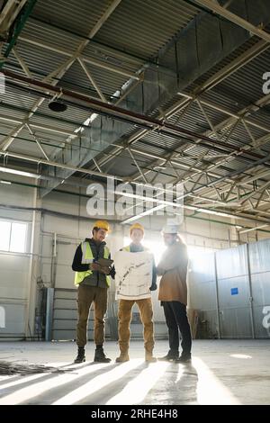 Long shot of three young engineers discussing building plan in blueprint at working meeting while standing next to each other in warehouse Stock Photo
