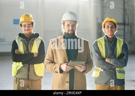Young successful foreman with tablet standing between two engineers in hardhats and protective fluorescent vests and looking at camera Stock Photo