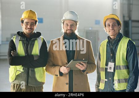 Three happy young intercultural engineers in protective helmets looking at camera with smiles while two men in safety vests surrounding foreman Stock Photo